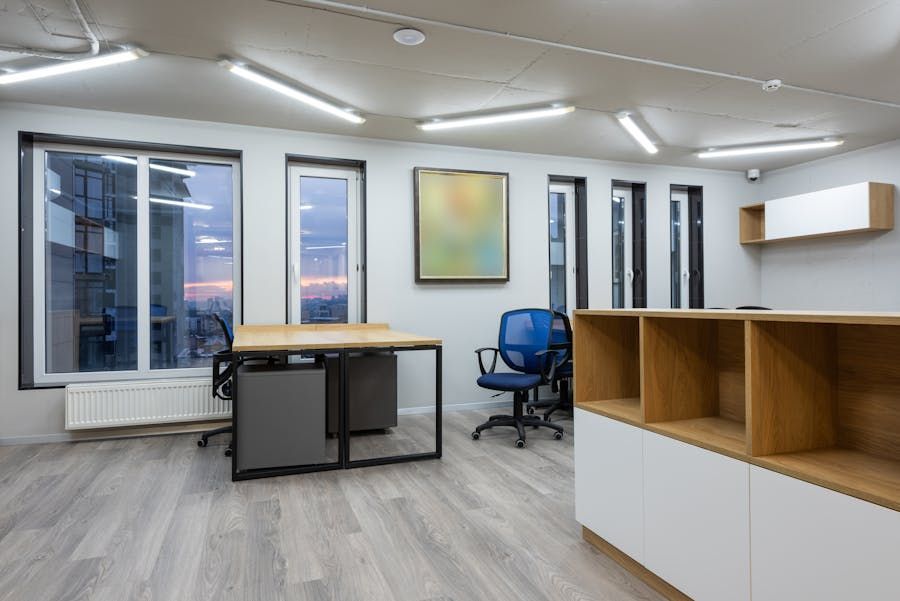 Modern office with desk, blue chair, open shelving, and large windows at dusk.