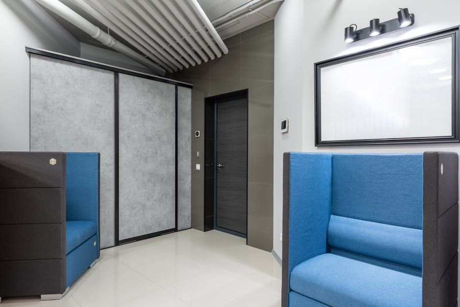 Small modern waiting room with blue chairs, gray walls, and a black-framed mirror under lights.