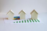 Three small white paper houses on a white surface, with a green-striped road in front.