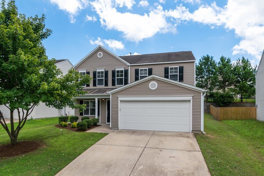 Two-story beige house with a white garage and driveway, front lawn, and tree under a blue sky