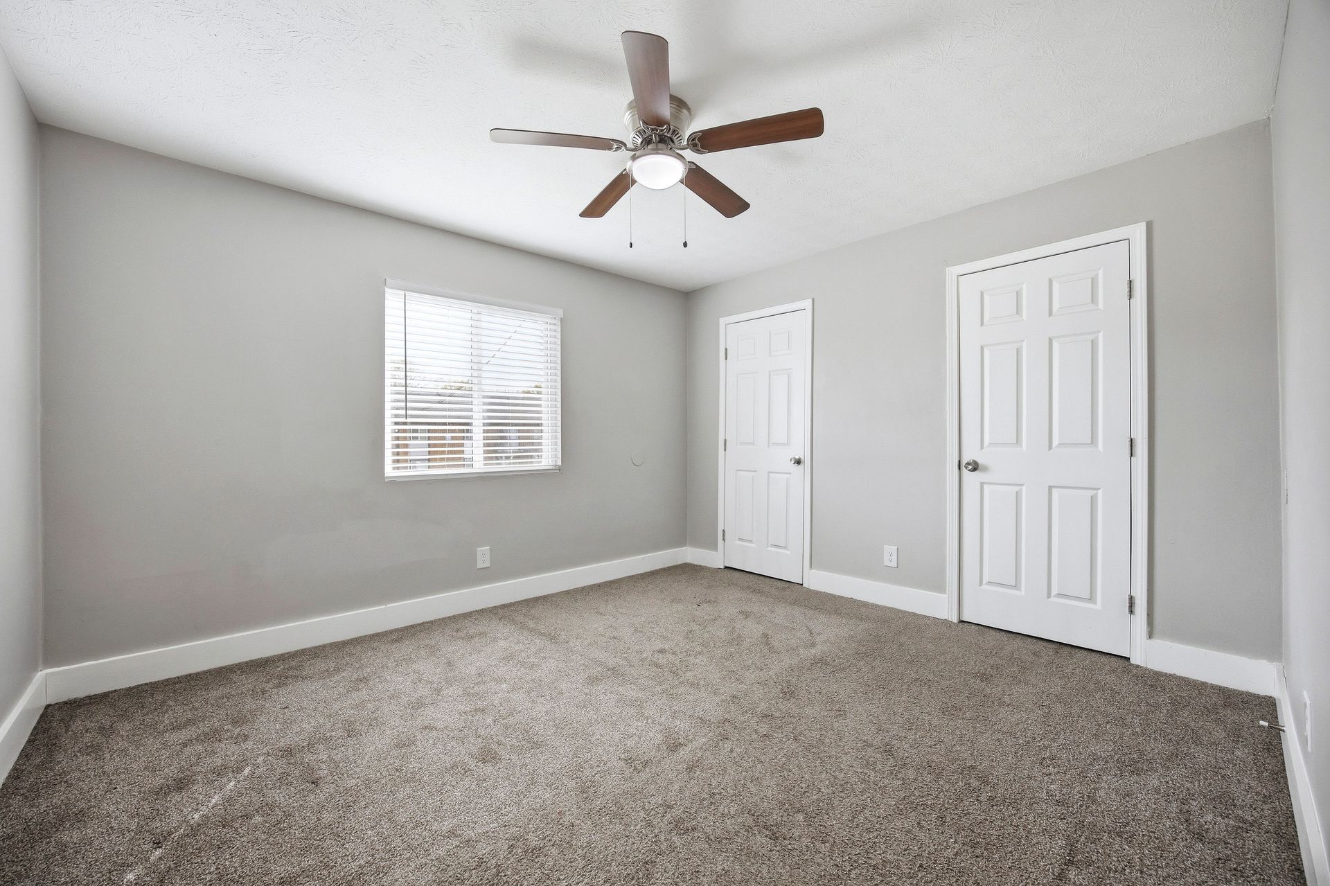 Empty gray bedroom with carpet, ceiling fan, window, and two white doors