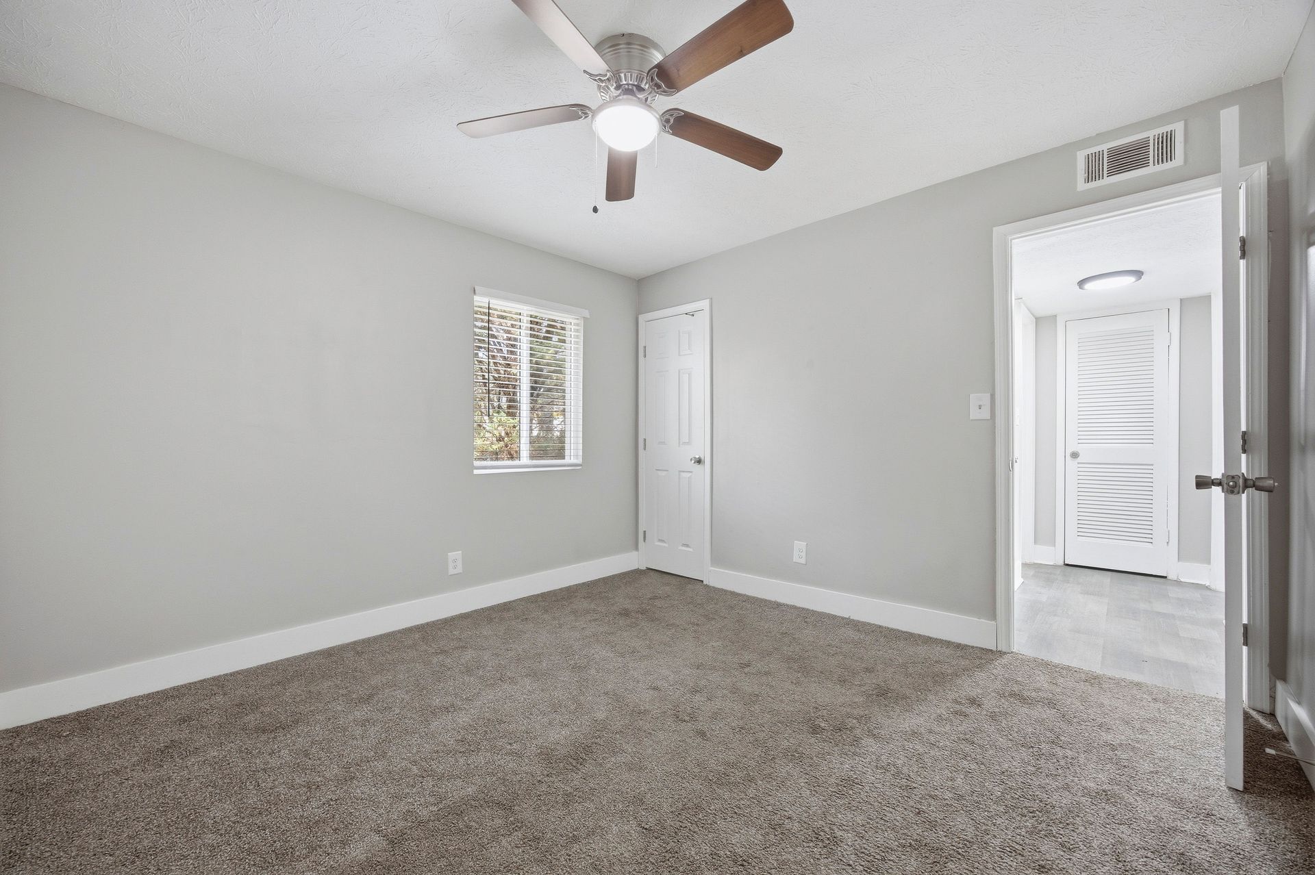 Empty gray bedroom with beige carpet, ceiling fan, window, closet doors, and an open doorway