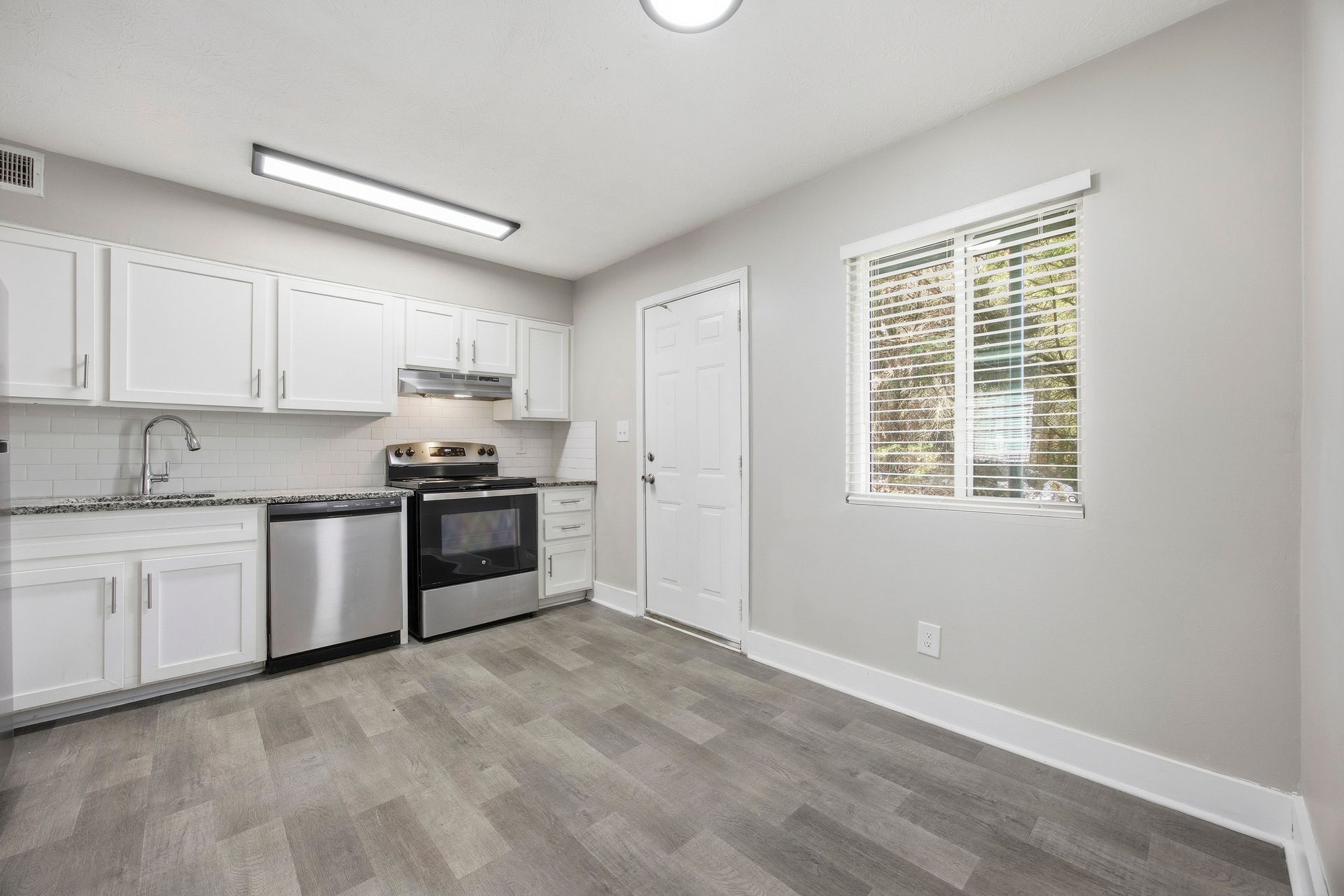 Empty white kitchen with stainless steel appliances, gray flooring, and a window letting in daylight