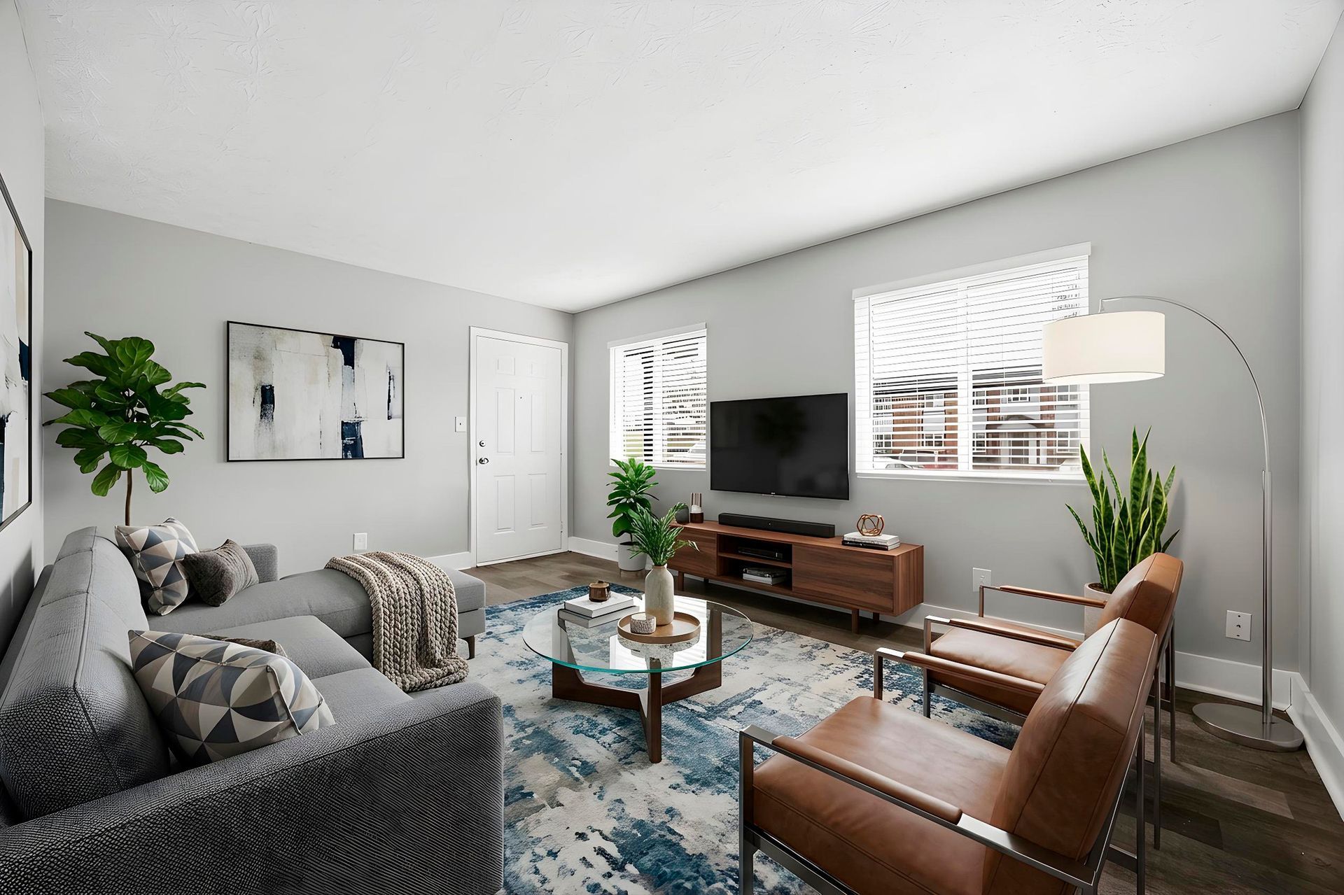 Bright living room with gray sofa, brown chairs, TV console, blue rug, and floor lamp by large windows