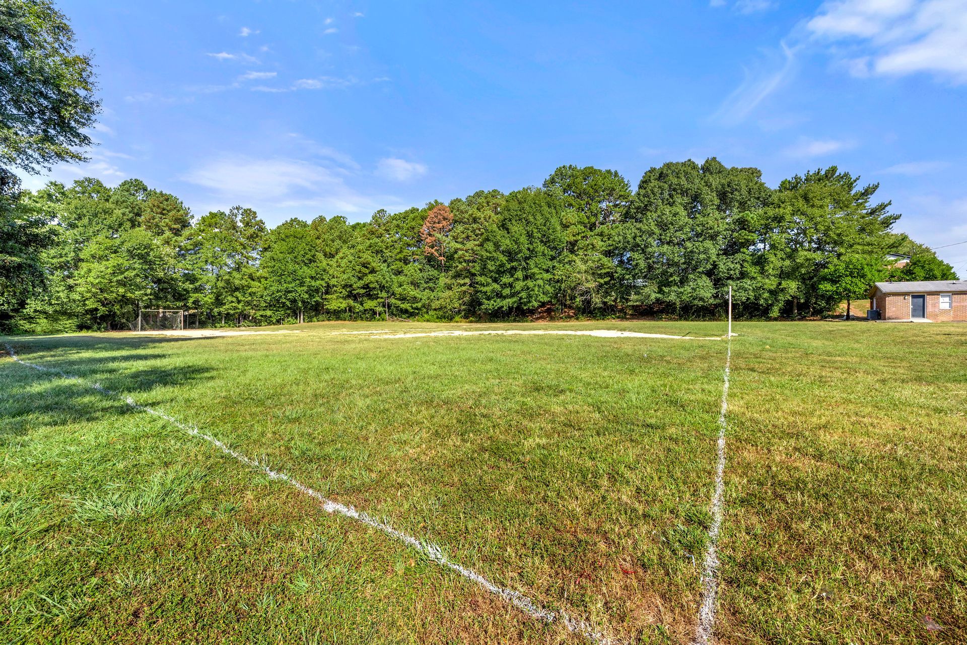 Grassy open field with white boundary lines, surrounded by trees under a blue sky, with a small building at right.