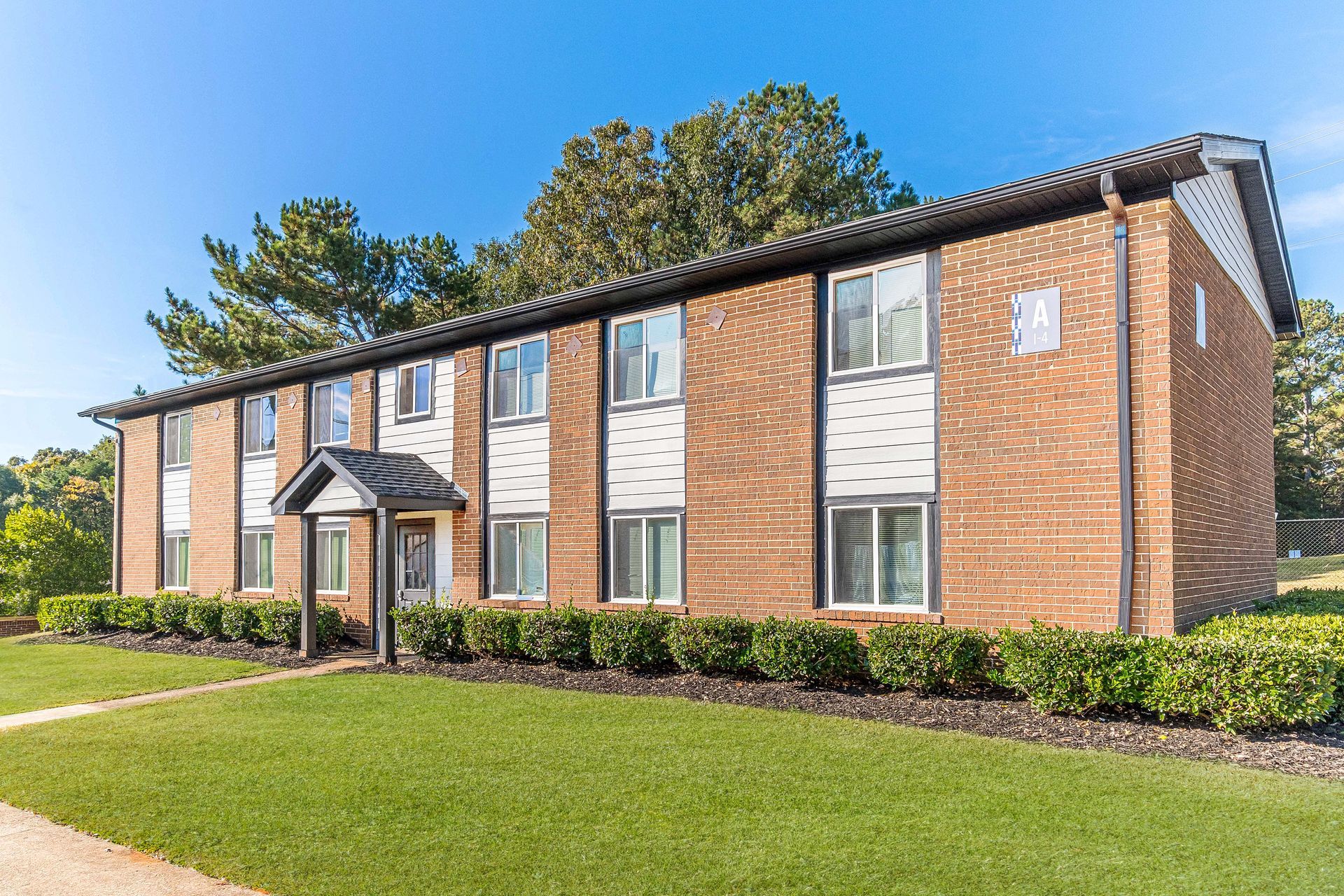 Two-story brick apartment building with white-trimmed windows, a small entrance awning, and green lawn under a blue sky
