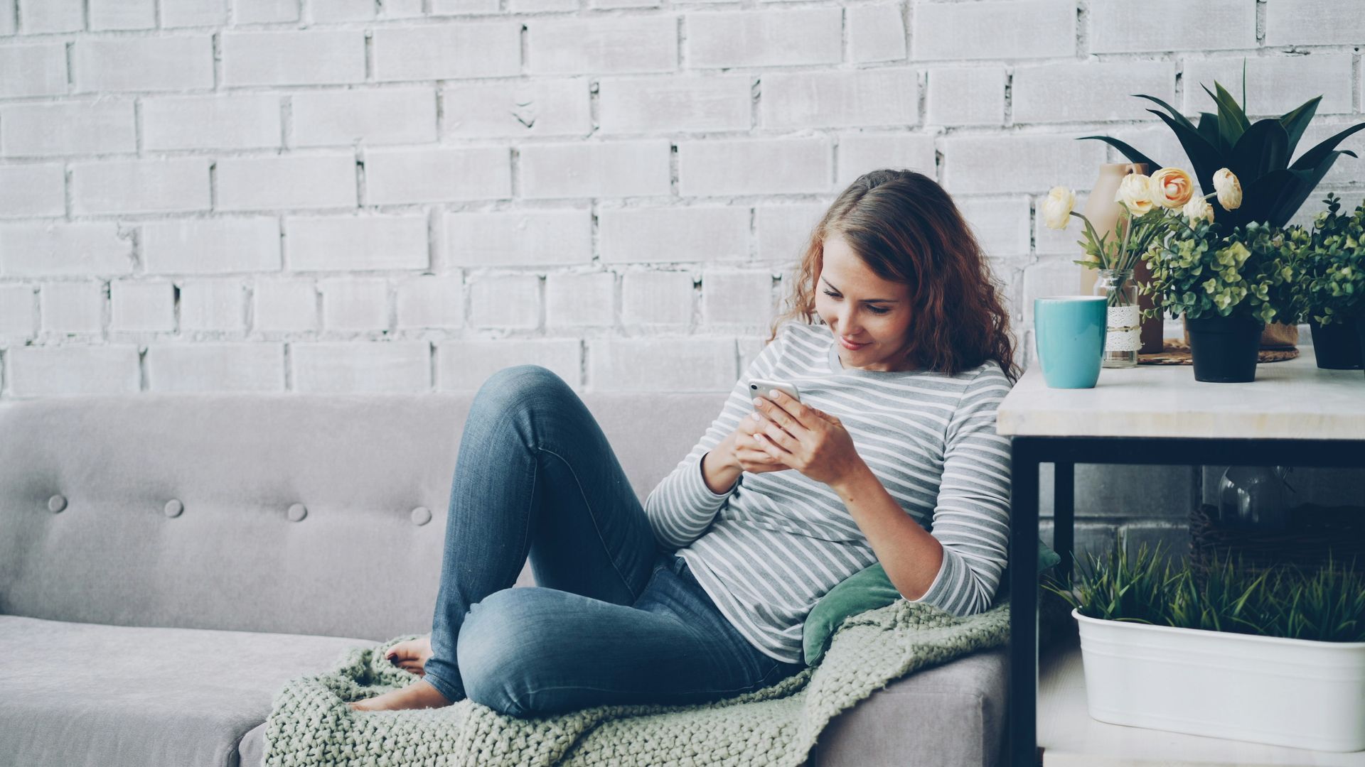 Woman sitting on a gray sofa, using a smartphone beside a table with flowers and a plant