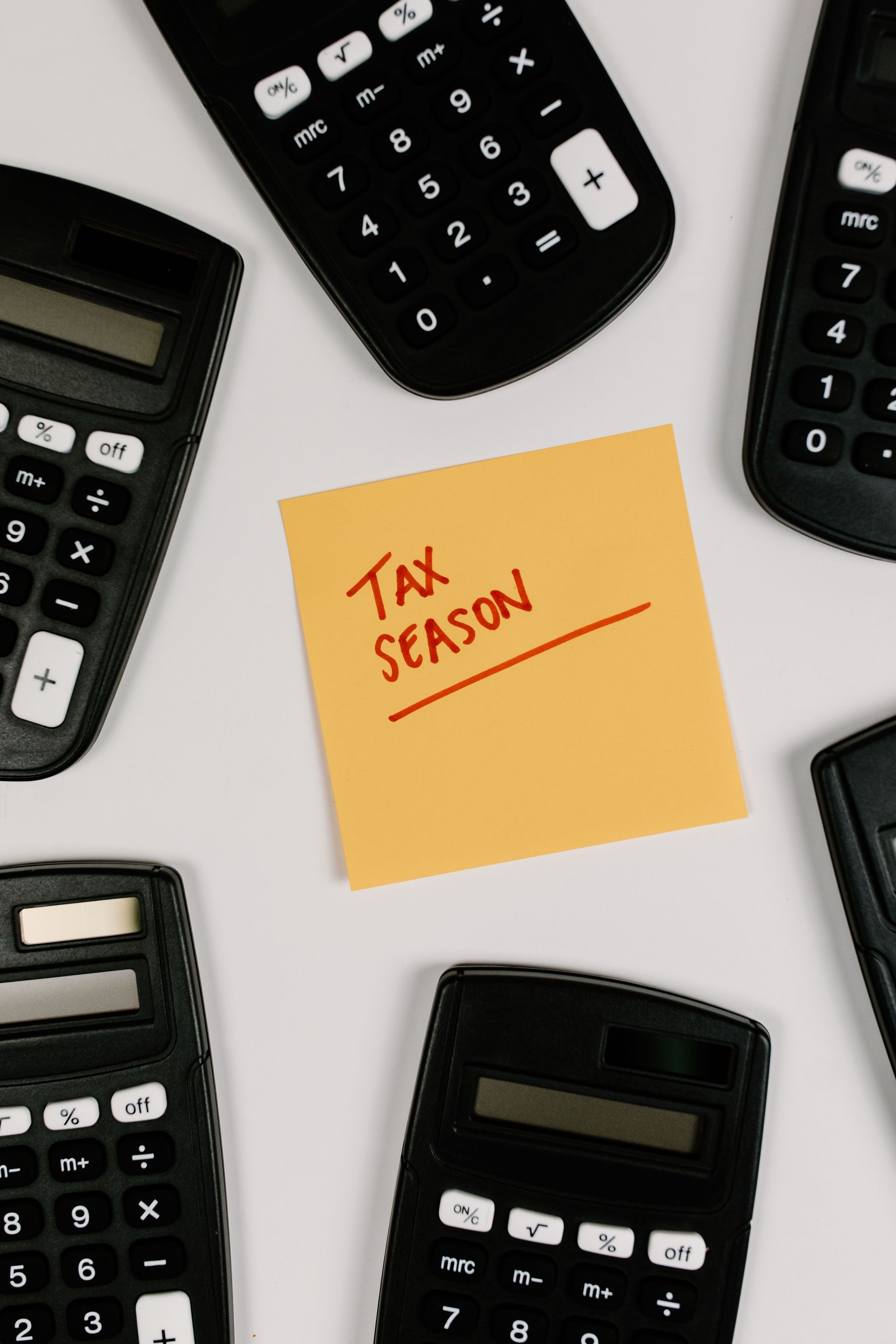 Yellow sticky note reading “Tax season” surrounded by calculators on a white background