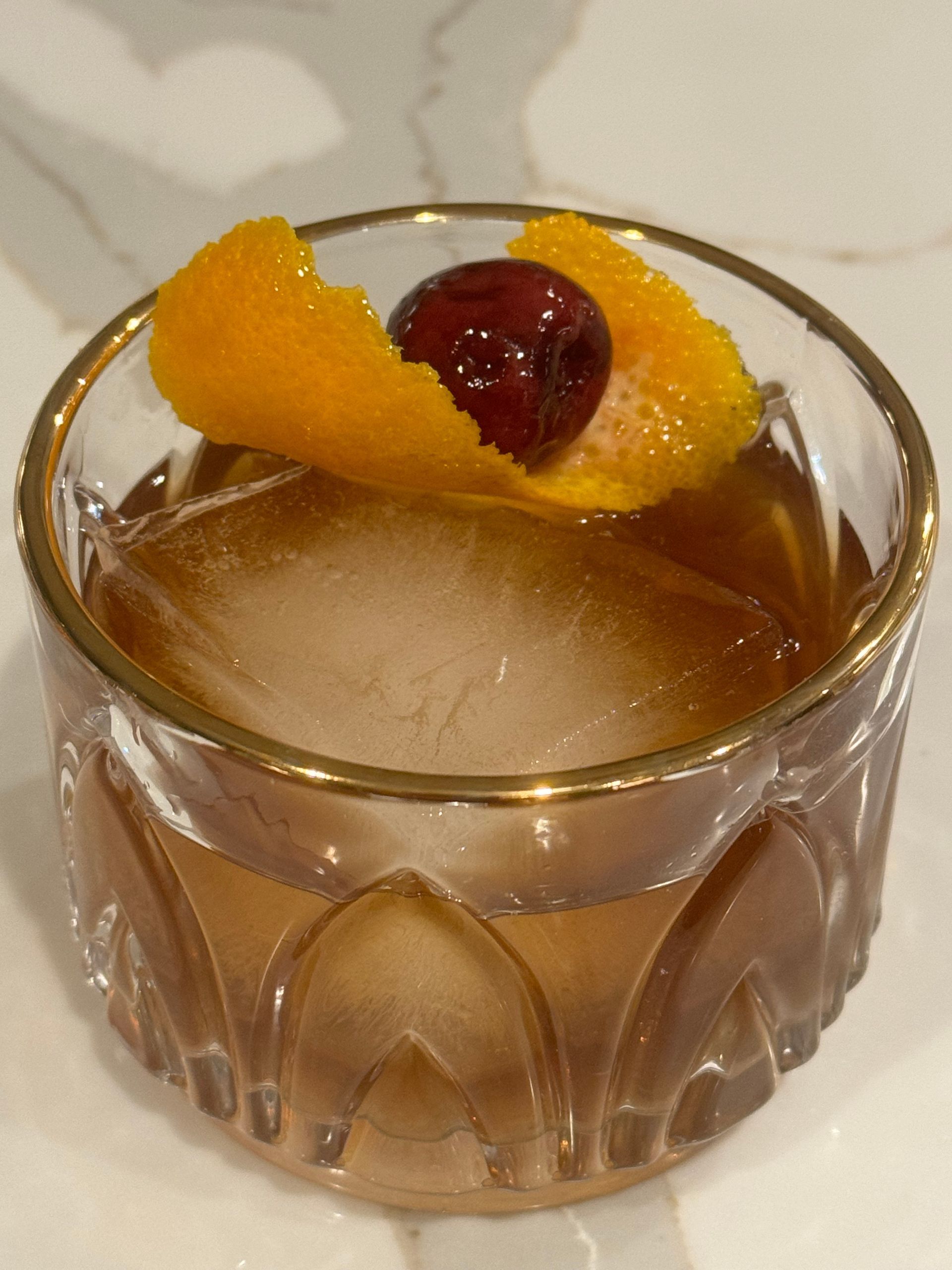 A glass filled with a drink with an orange peel and a cherry on top.