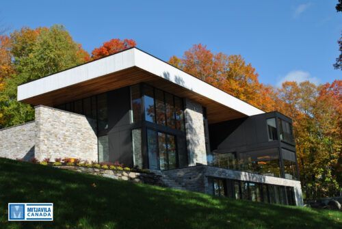 Maison moderne aux murs de pierre, fa&ccedil;ade vitr&eacute;e et toit plat, adoss&eacute;e &agrave; une colline orn&eacute;e d'arbres aux couleurs automnales.
