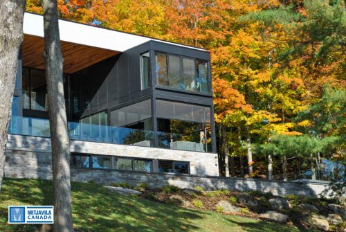 Maison moderne noire aux murs de verre, nich&eacute;e au milieu d'arbres aux feuillages d'automne flamboyants.