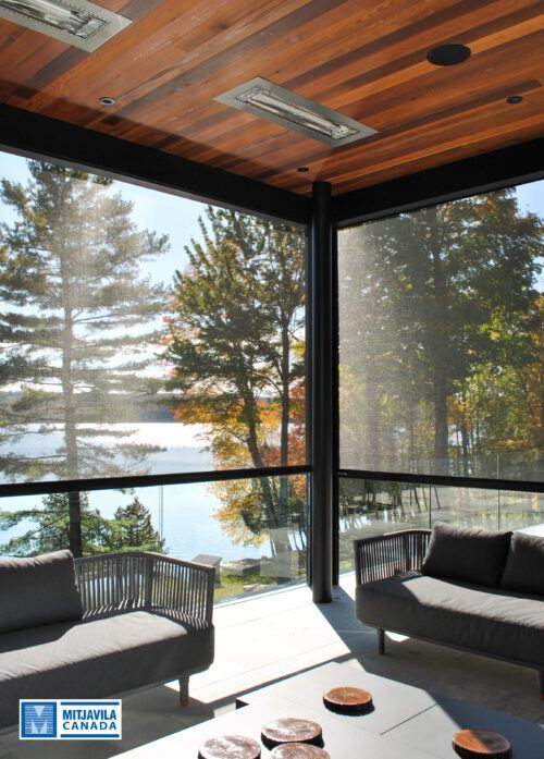 V&eacute;randa avec vue sur le lac, mobilier gris, plafond en bois et arbres visibles.