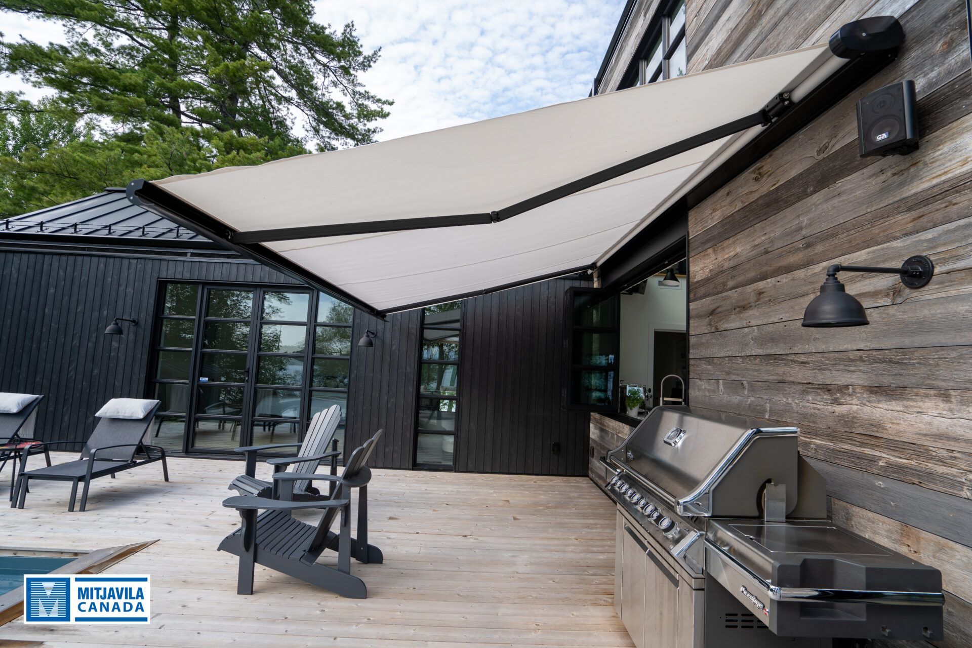 Terrasse extérieure avec auvent ouvert, barbecue et sièges. Bardage en bois et bâtiment noir en arrière-plan.
