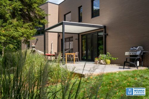Terrasse avec pergola, table, barbecue et hautes herbes dans un jardin jouxtant un b&acirc;timent moderne.