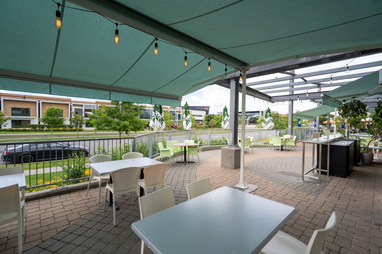 Terrasse extérieure de restaurant donnant sur un étang. Tables et chaises sous un auvent turquoise, guirlandes lumineuses.
