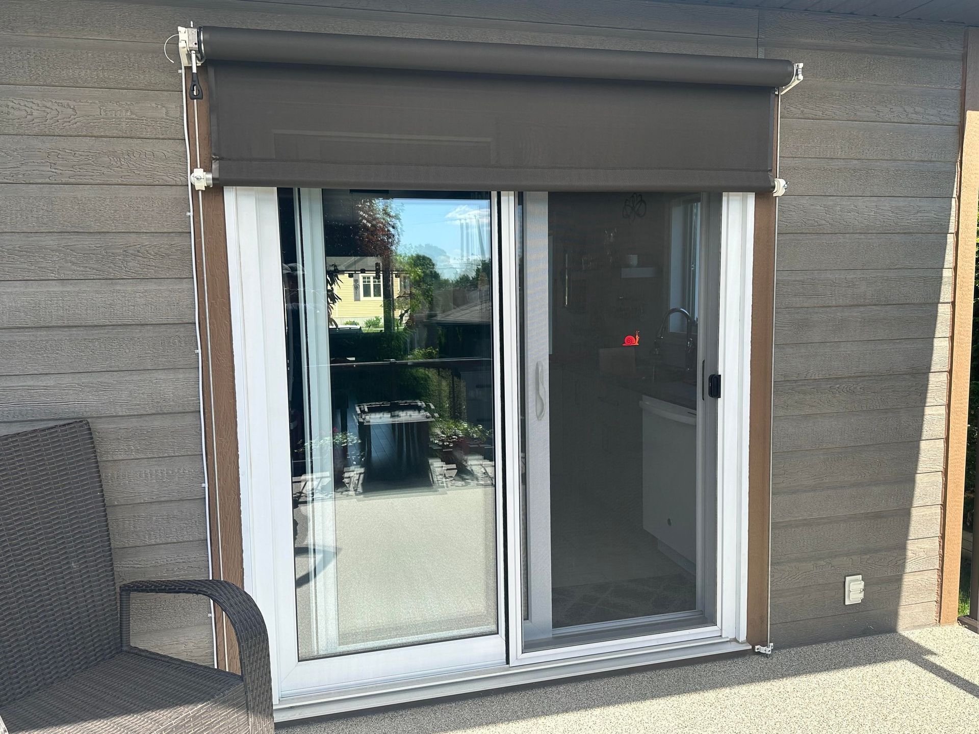 Porte coulissante en verre avec store marron, encadrée par un revêtement en pierre. Une chaise de patio se trouve à proximité.