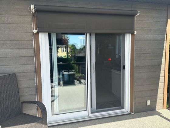 Porte coulissante en verre avec store marron, encadrée par un revêtement en pierre. Une chaise de patio se trouve à proximité.