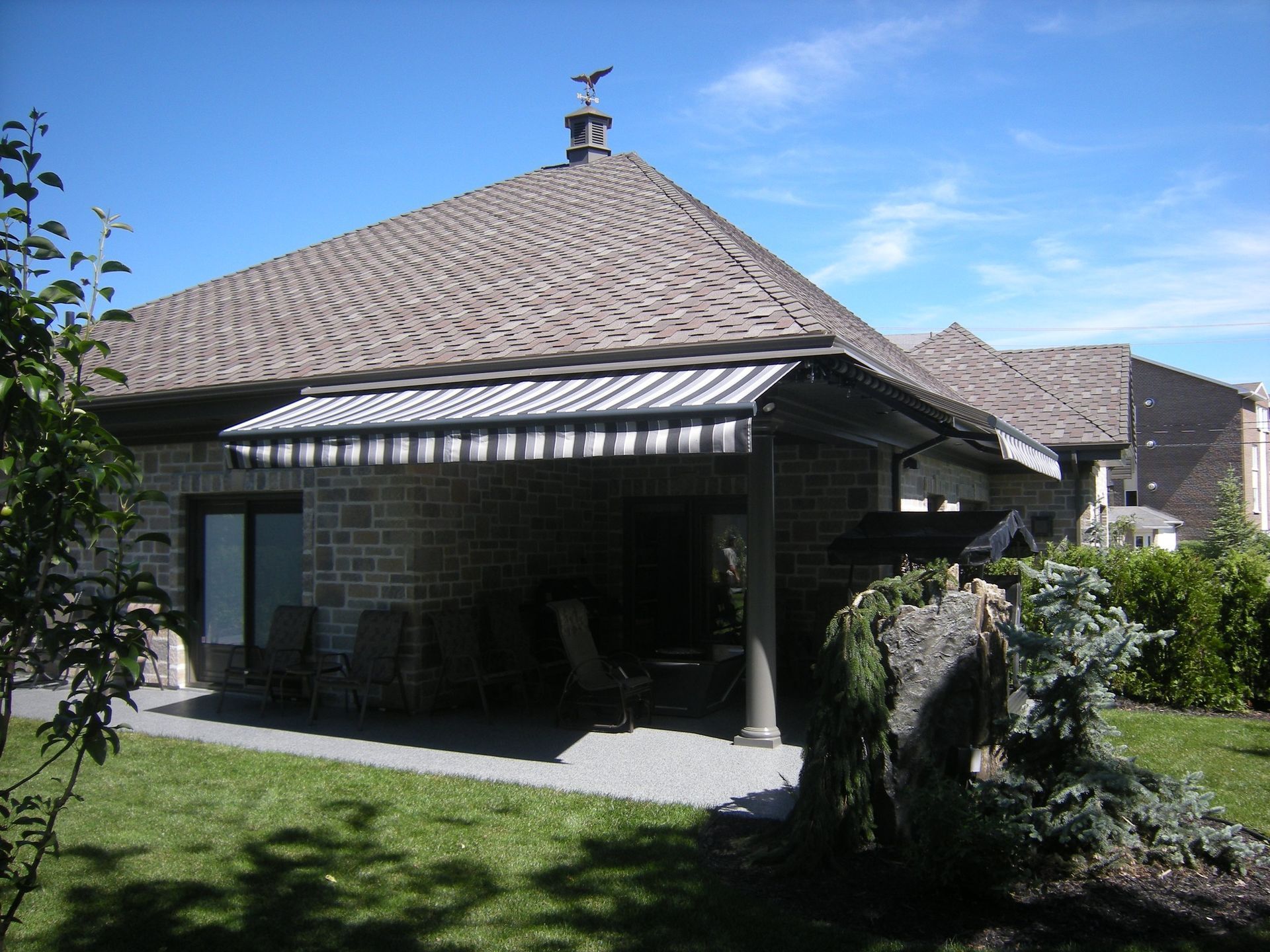 Maison en pierre avec auvent au-dessus de la terrasse, pelouse verte, ciel bleu.