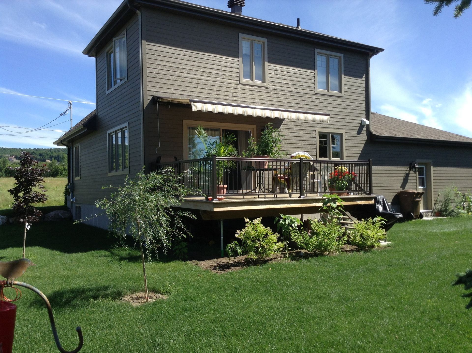 Maison à deux étages avec terrasse en bois. Pelouse verte, ciel bleu. La terrasse est aménagée avec des plantes, du mobilier et un auvent.