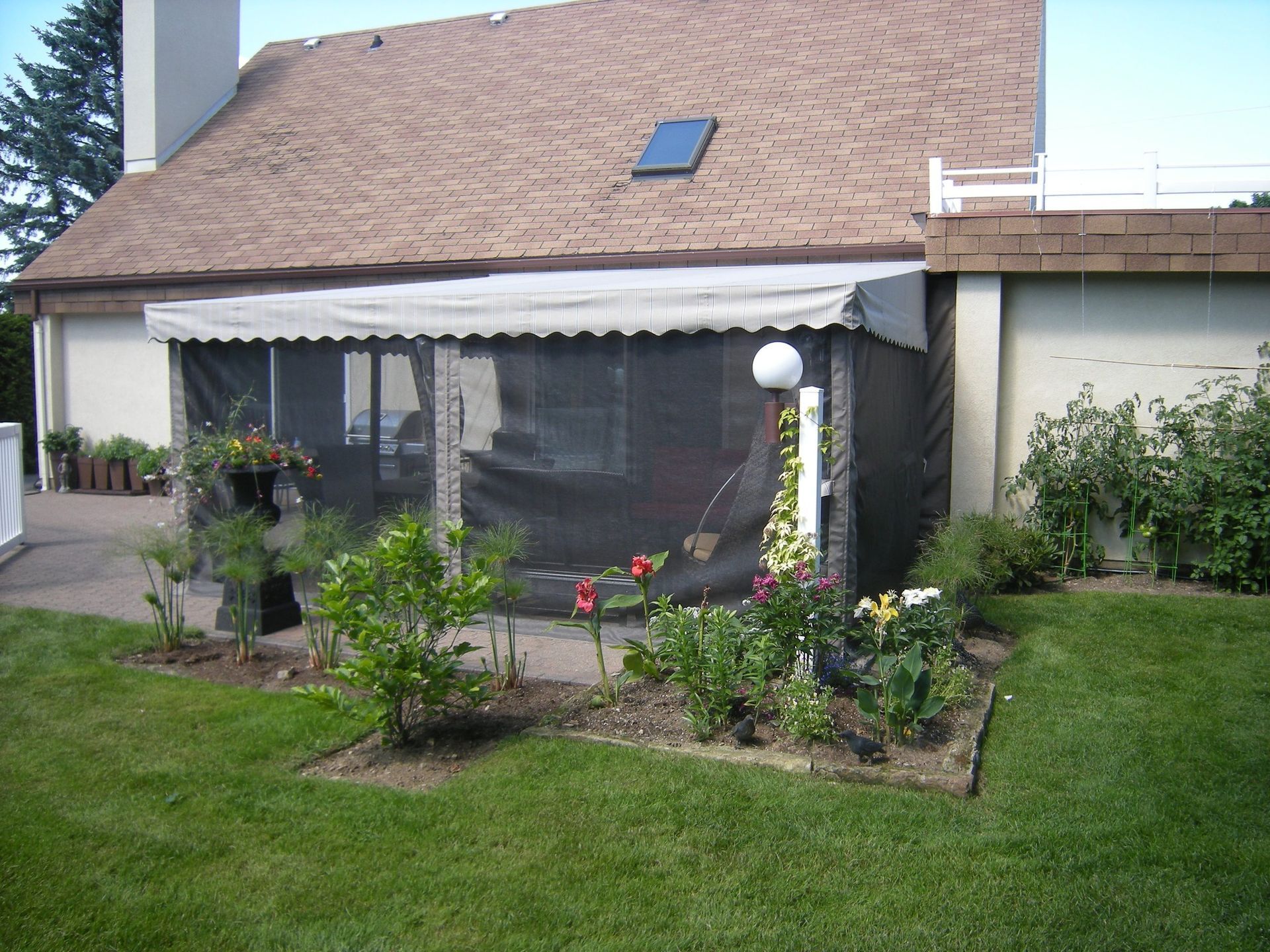 Véranda avec auvent, attenante à une maison. Jardin fleuri et gazonné devant.