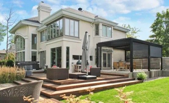 Vue arrière d'une maison en stuc clair avec une terrasse en bois, des sièges extérieurs et une pergola.
