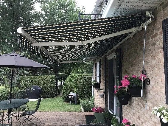 Un auvent à rayures noires et blanches s'étend au-dessus d'une porte-fenêtre. Des fleurs roses en pots suspendus sont visibles sur le mur de briques.
