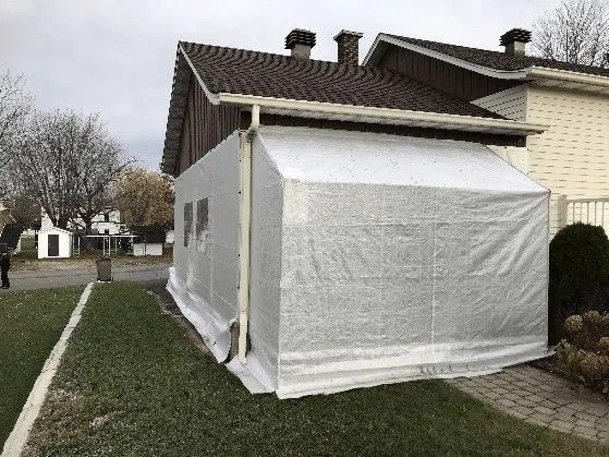 Une structure recouverte de plastique blanc, adossée à une maison. Vue extérieure, sur l'herbe, avec gouttière et bâtiment en briques visibles.