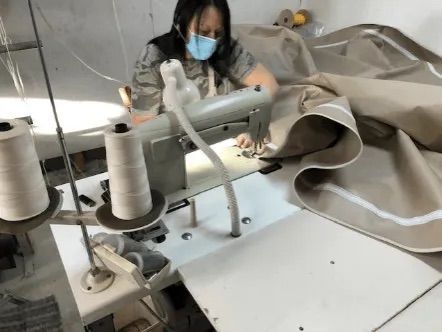 Femme portant un masque utilisant une machine à coudre industrielle pour coudre un tissu beige à bordure blanche dans un environnement d'usine.