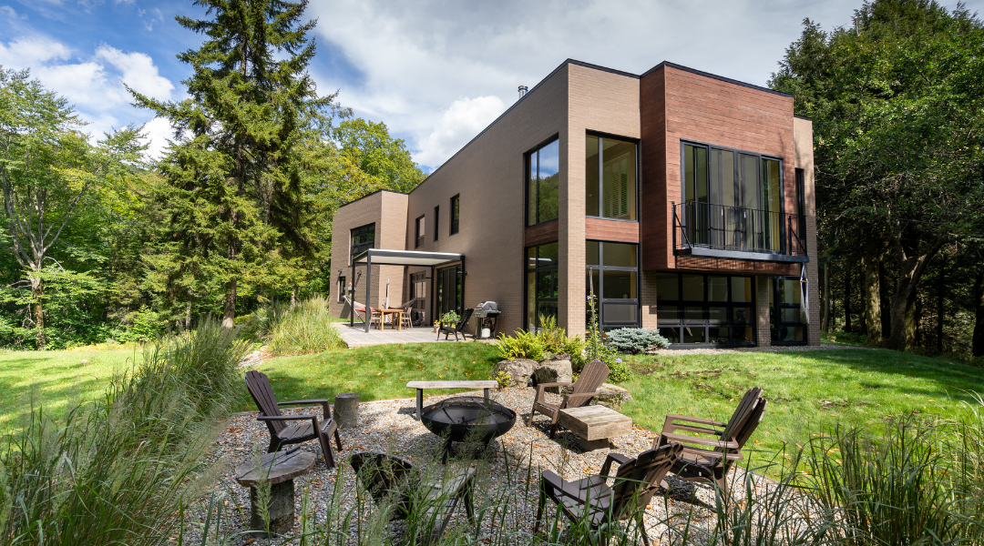Maison moderne à l'extérieur en briques brunes, située dans un paysage naturel avec un petit étang à l'avant.