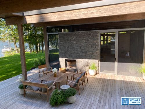 Terrasse ext&eacute;rieure avec plancher en bois, mobilier de jardin, chemin&eacute;e, donnant sur un lac.
