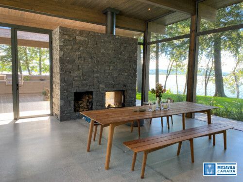 Salle &agrave; manger avec table et banc en bois, chemin&eacute;e en pierre et grandes fen&ecirc;tres donnant sur un lac.