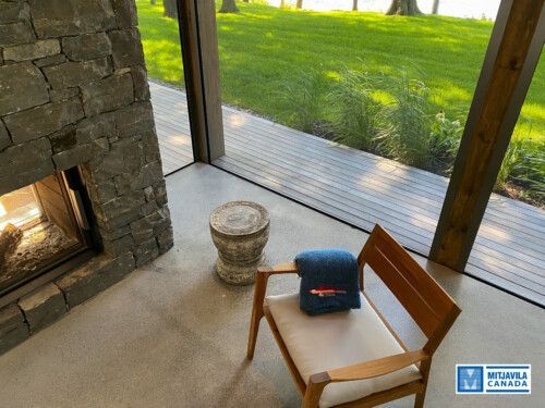 Chemin&eacute;e en pierre, fauteuil en bois recouvert d'une couverture bleue, sol en b&eacute;ton et grande fen&ecirc;tre donnant sur la verdure.