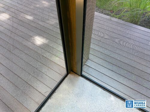 Coin d'un cadre de porte moustiquaire; rencontre entre une terrasse grise et un sol blanc.