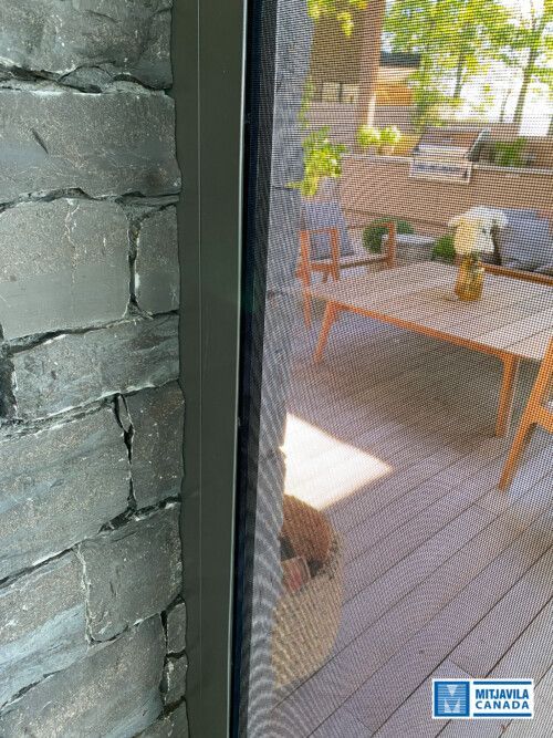 Gros plan sur une porte moustiquaire adoss&eacute;e &agrave; un mur de pierre. Un chien est visible &agrave; travers la moustiquaire, regardant vers l'ext&eacute;rieur.