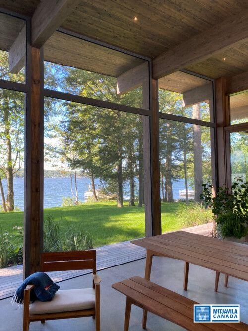 Terrasse au bord du lac avec mobilier en bois et grandes fen&ecirc;tres donnant sur le lac et les arbres.