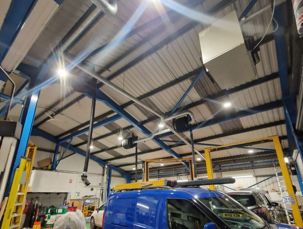 Inside of a brightly lit industrial vehicle workshop with a blue van parked beneath overhead ventilation pipes.