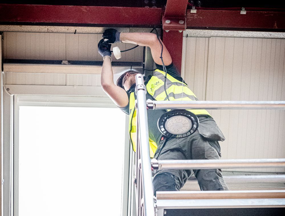 A construction worker in a high-visibility vest stands on scaffolding, working on electrical wiring near a ceiling.