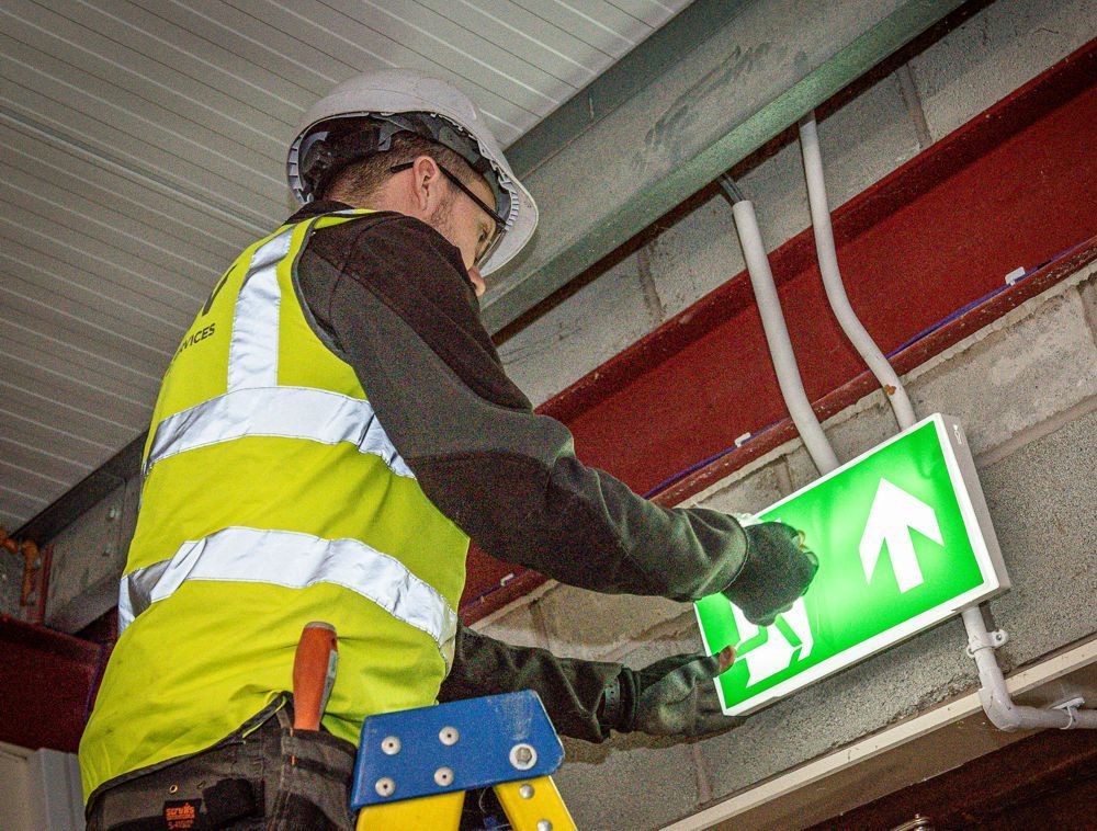 A worker in a high-visibility vest and hard hat installing an illuminated green exit sign on an industrial ceiling.