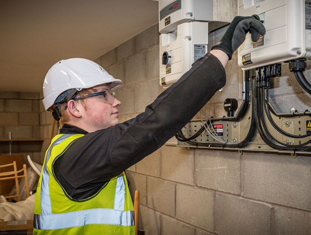 A technician in a hard hat and high-visibility vest inspects electrical equipment mounted on a grey cinder block wall.