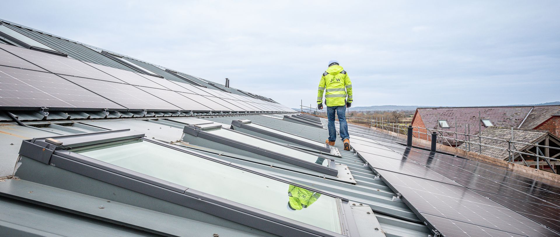A worker in a bright yellow high-visibility jacket walks across a large, slanted roof fitted with skylights and solar panels.