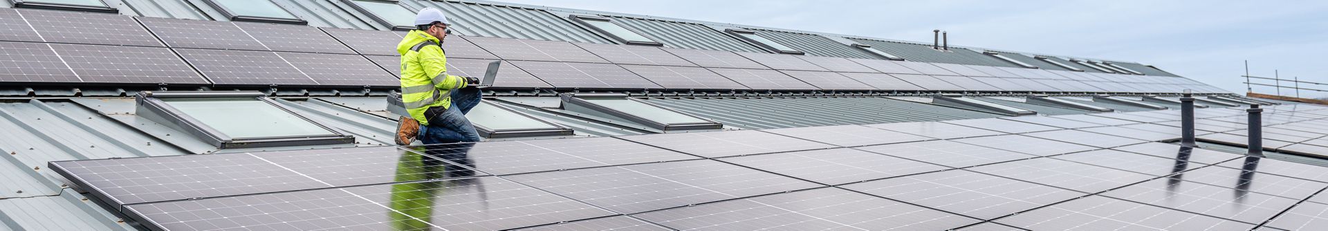 Worker in a neon jacket on a rooftop lined with solar panels, inspecting or cleaning them under an overcast sky