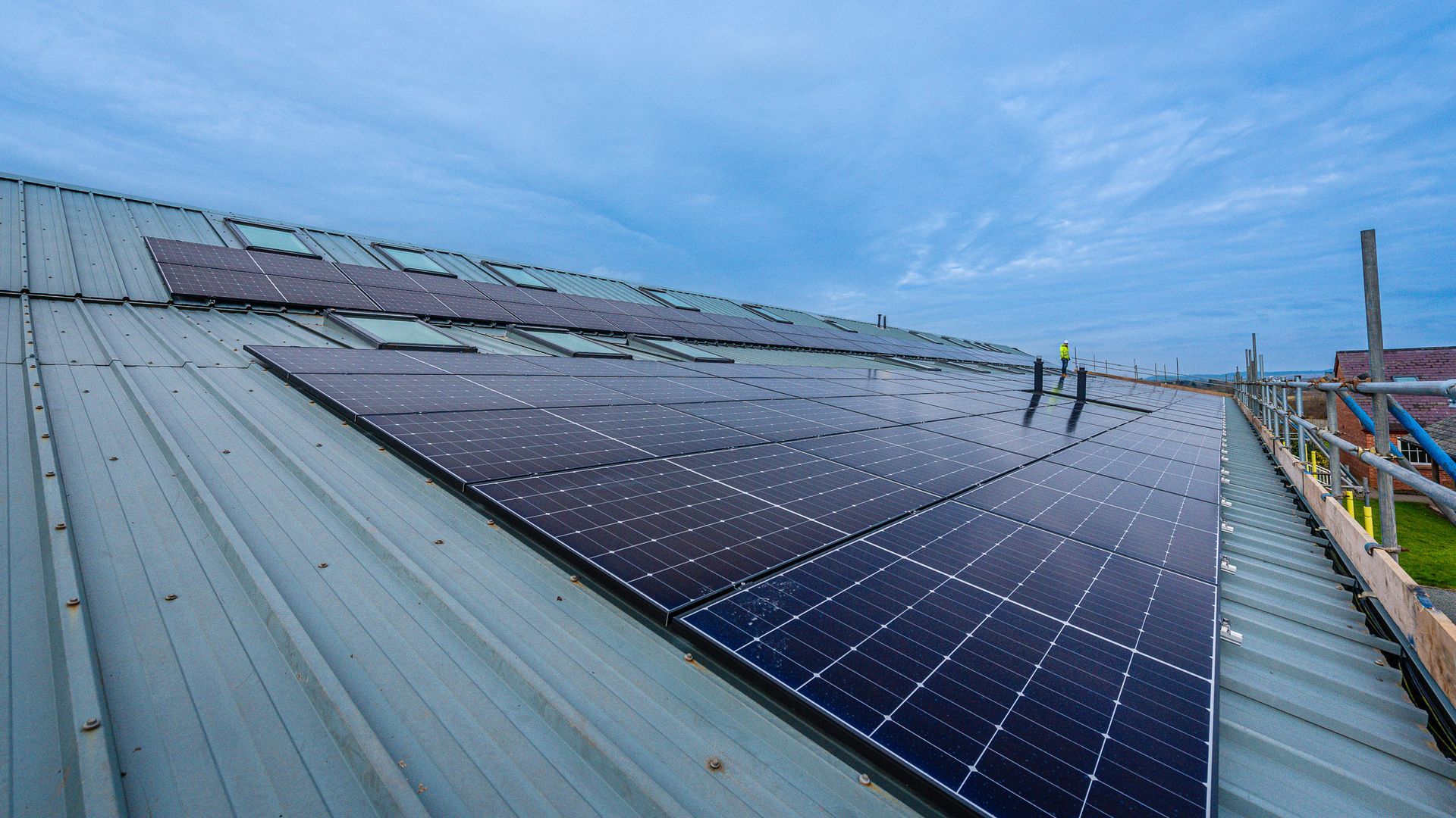 A large array of black solar panels installed on a sloped metal roof under a blue sky, with scaffolding on the side.