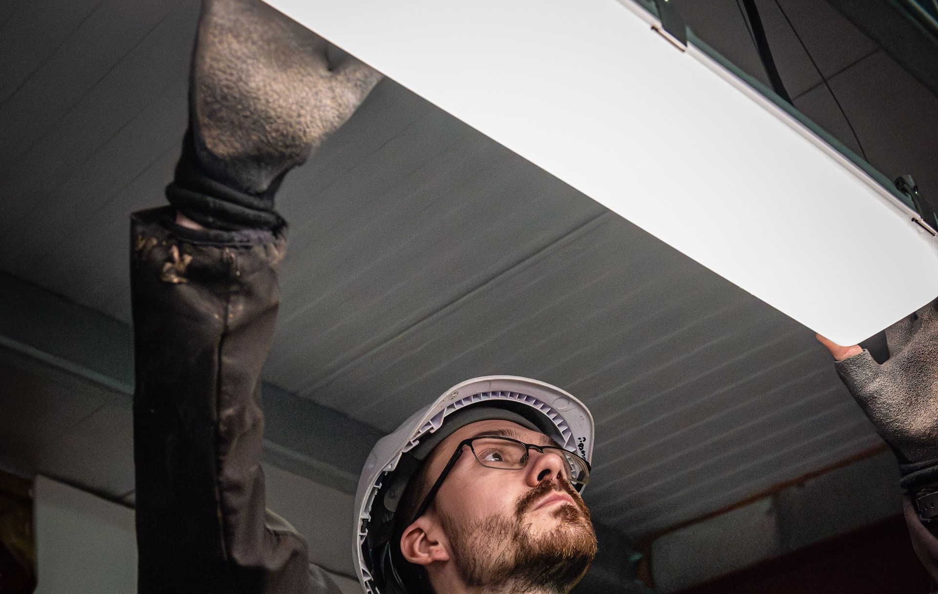 A person wearing a hard hat and gloves installs a long, bright rectangular ceiling light fixture.