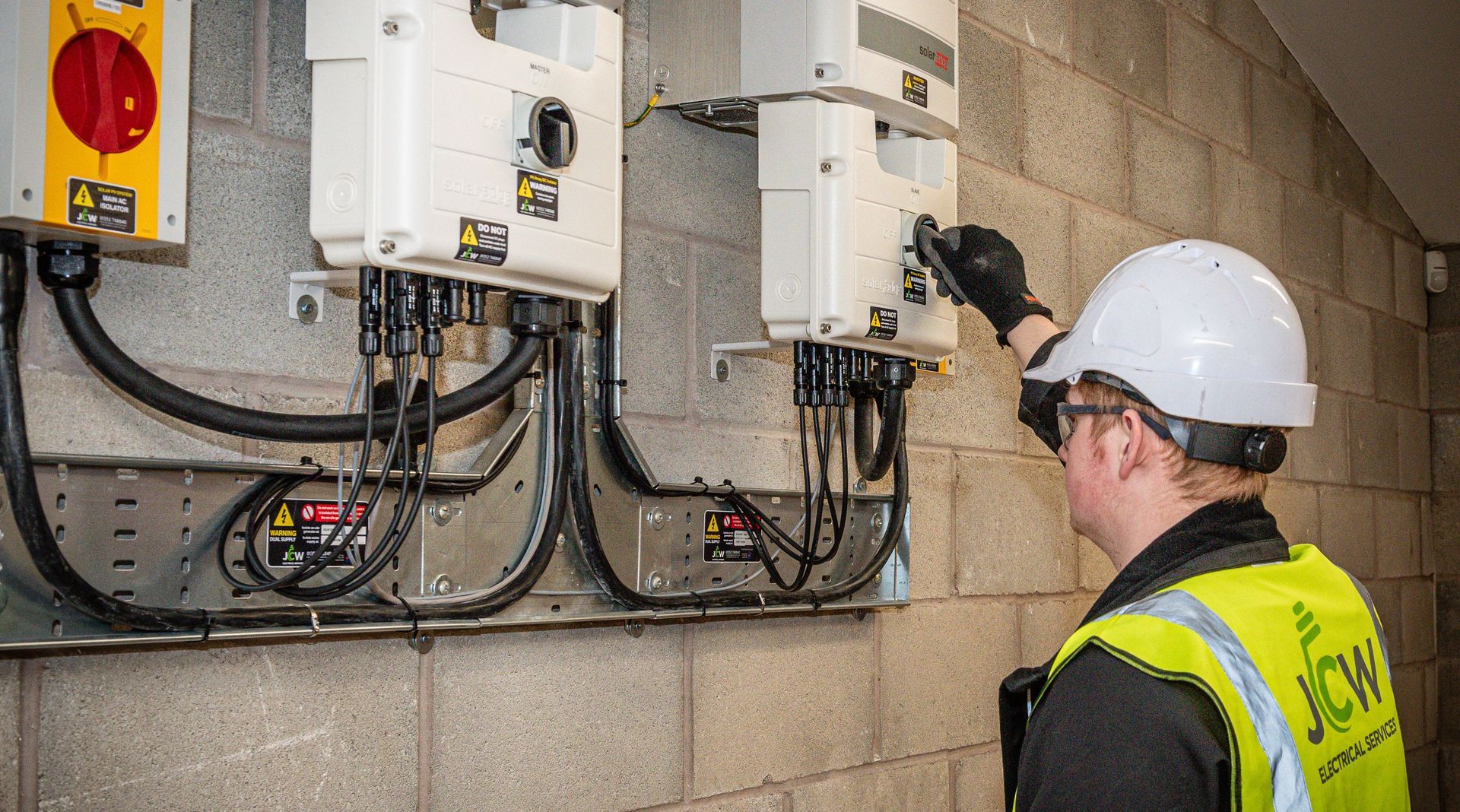 A technician in a hard hat and safety vest performs maintenance on industrial wall-mounted electrical equipment.