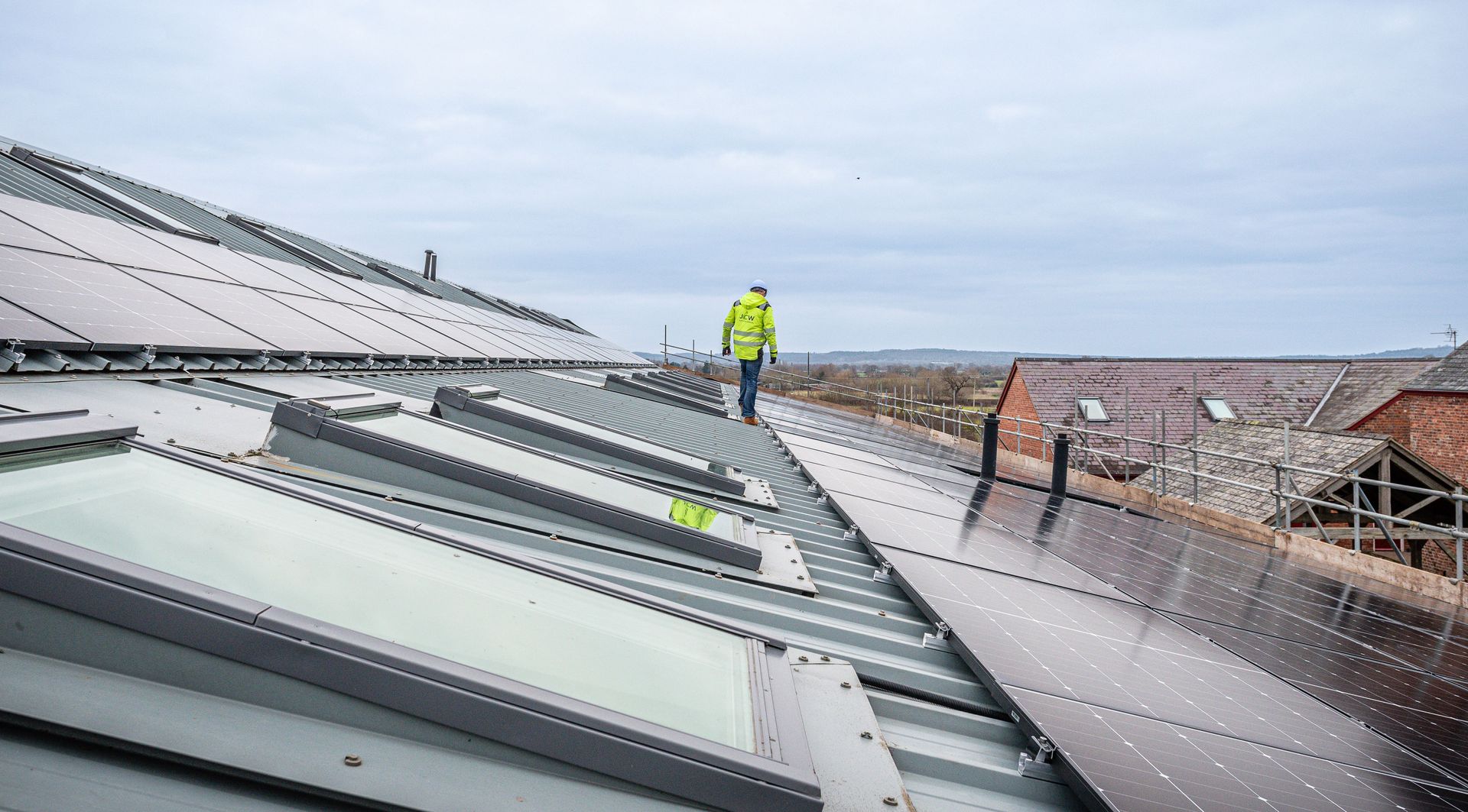 A worker in a high-visibility jacket walks along a sloped industrial roof featuring solar panels and skylights.