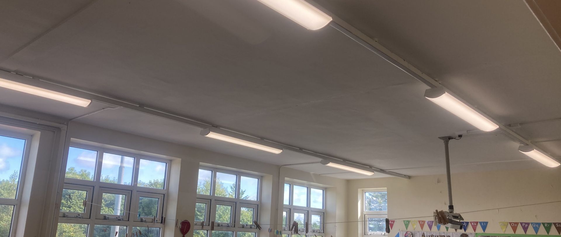 Interior view of a classroom ceiling with long, rectangular light fixtures and a ceiling-mounted projector.