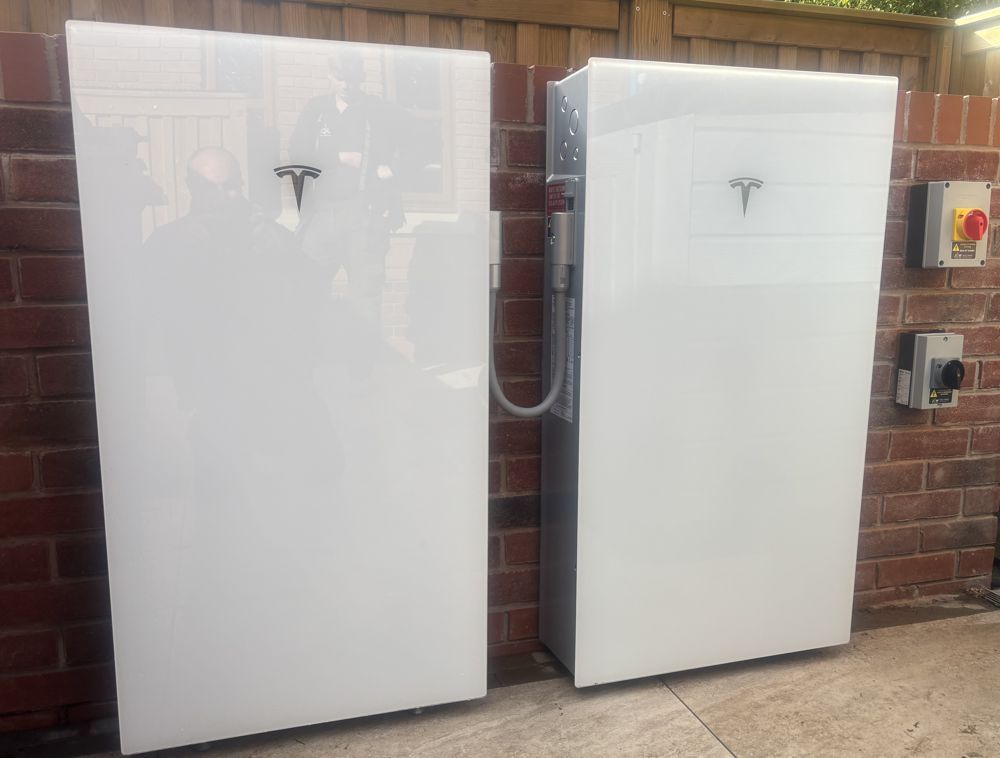 Two white, rectangular Tesla Powerwall home battery units mounted on a brick wall next to electrical switches.