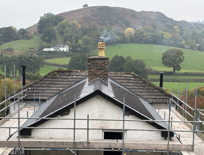 A roof under construction with scaffolding, a smoking brick chimney, and a rural hillside background.
