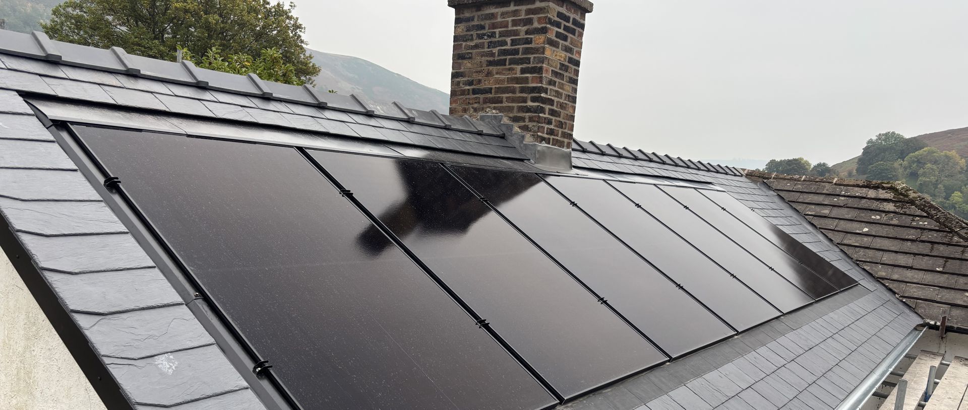 A row of sleek black solar panels integrated into a slate-tiled roof next to a brick chimney.