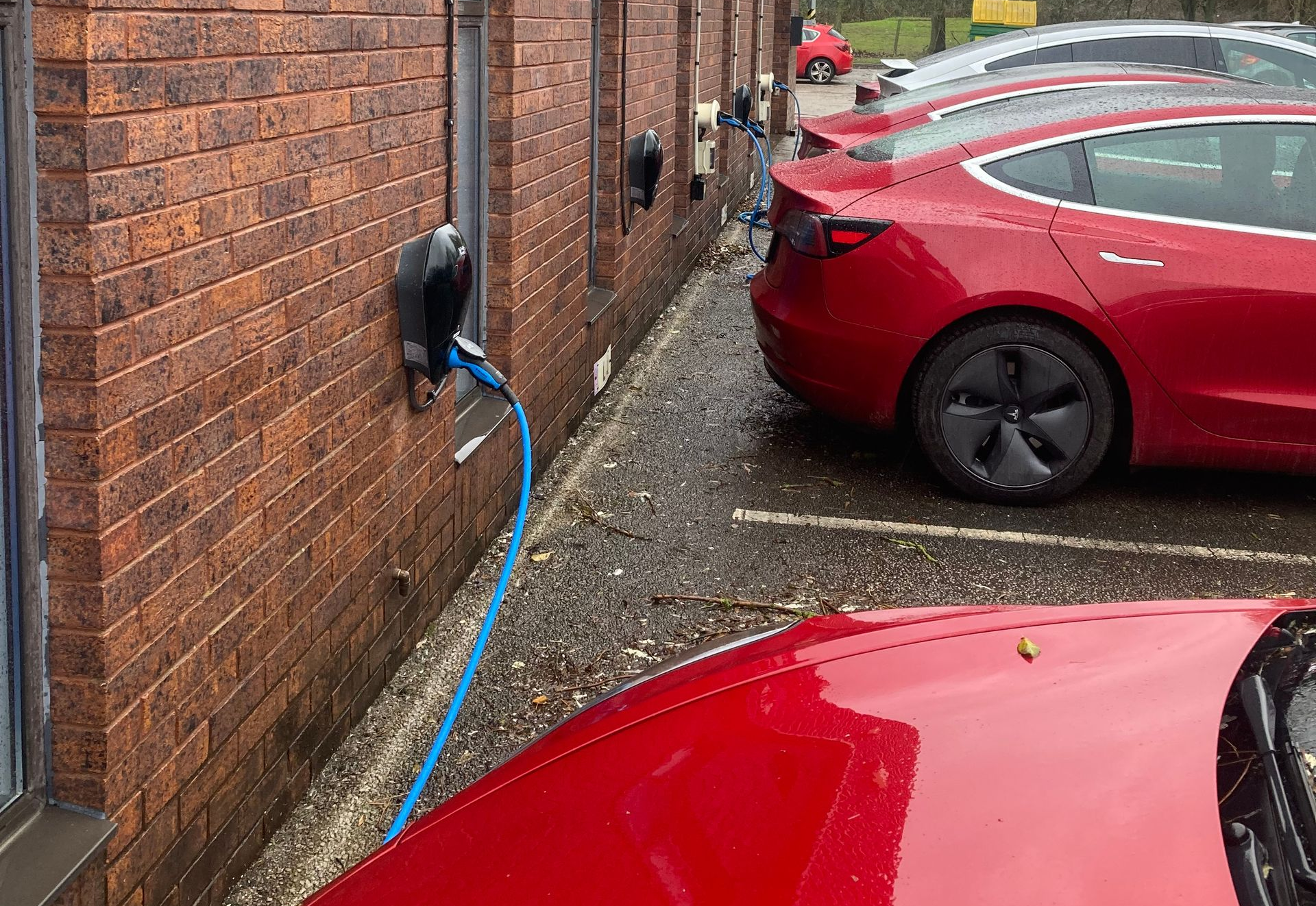 Two red Tesla electric vehicles parked side-by-side in a brick wall lot, charging via blue cables from the wall.